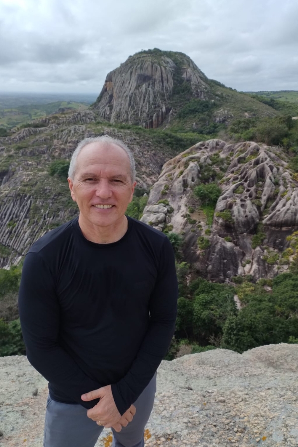 Um homem sorridente de camiseta preta posa em primeiro plano. Ao fundo, destaca-se uma imensa formação rochosa vertical e arredondada cercada por colinas e vegetação, sob um céu encoberto no Parque Estadual Pedra da Boca.