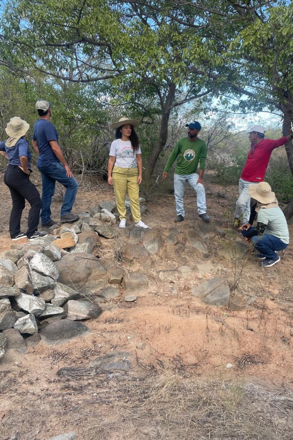 Grupo de seis pessoas com chapéus reunido em uma área de caatinga, conversando ao lado de uma estrutura de contenção feita com pedras empilhadas.