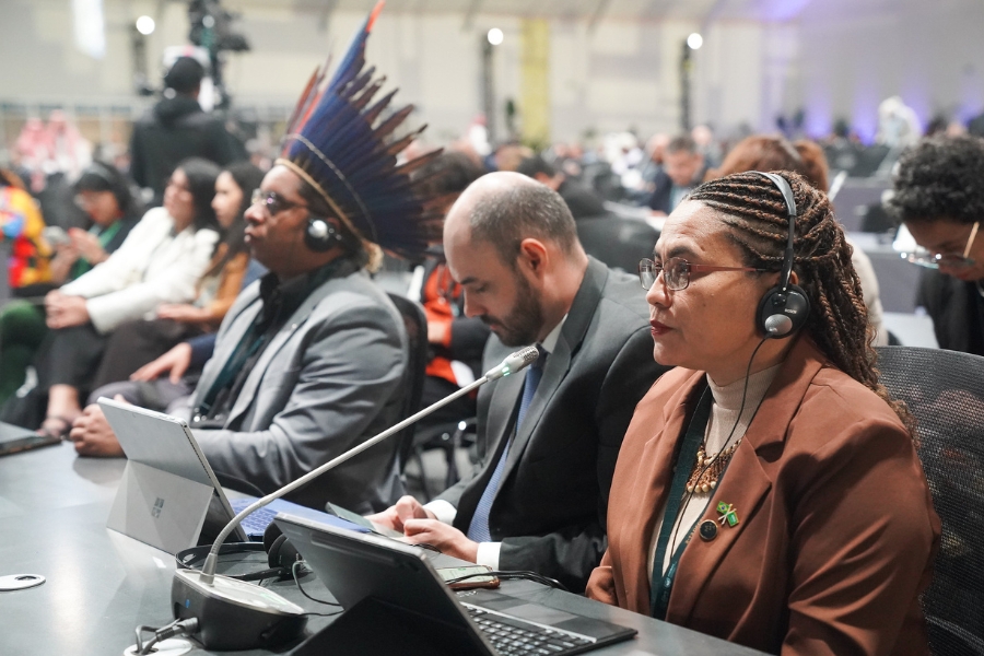 Delegação sentada em uma mesa de conferência internacional. Em primeiro plano, uma mulher de casaco marrom e fones de ouvido; ao fundo, um homem com cocar indígena tradicional.