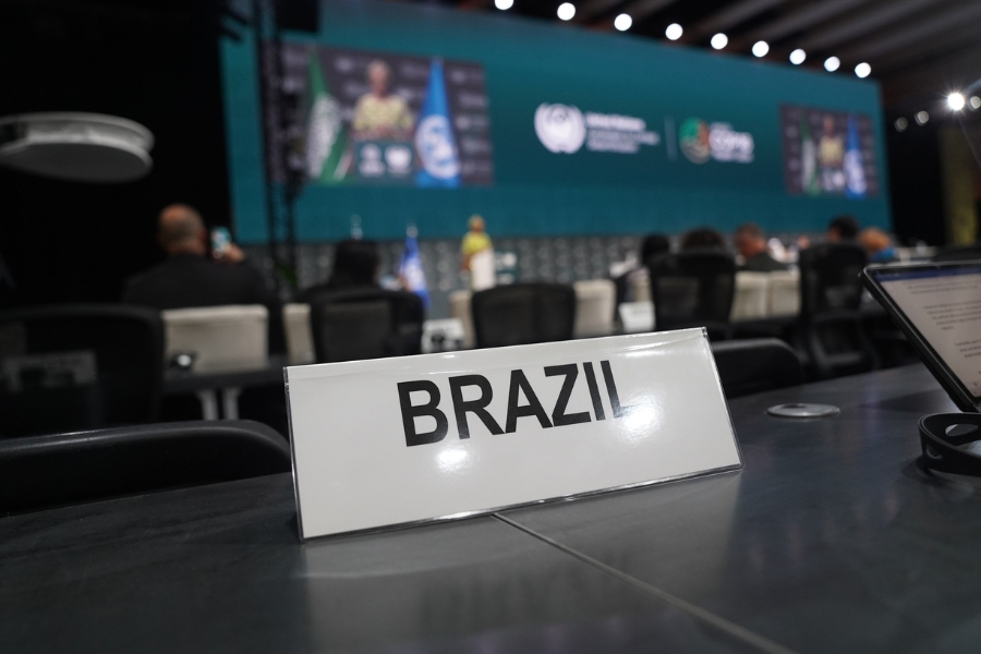 Placa branca com a palavra "BRAZIL" em letras pretas maiúsculas, posicionada sobre uma mesa preta. Ao fundo desfocado, vê-se um grande telão azul em um auditório de conferência.