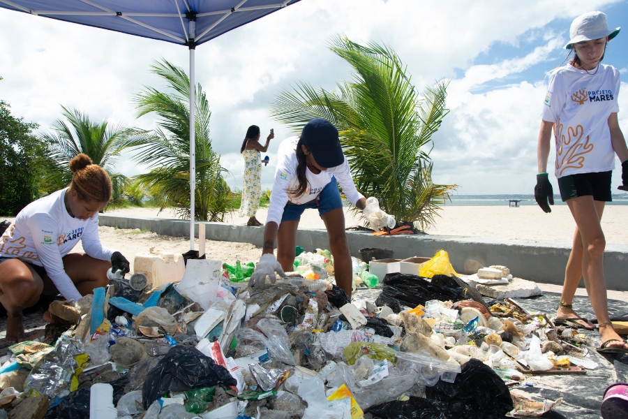 Três voluntários do "Projeto Mares" realizam a limpeza de uma praia. Eles estão sob uma tenda, separando uma grande pilha de resíduos plásticos, garrafas de vidro e entulhos coletados da areia.