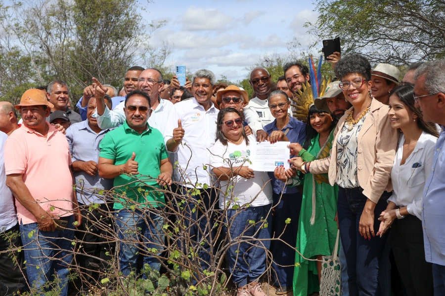 Grande grupo de pessoas sorrindo e posando ao ar livre atrás de um pequeno arbusto. Algumas pessoas seguram um documento impresso no centro, cercadas por autoridades e uma pessoa com cocar indígena.