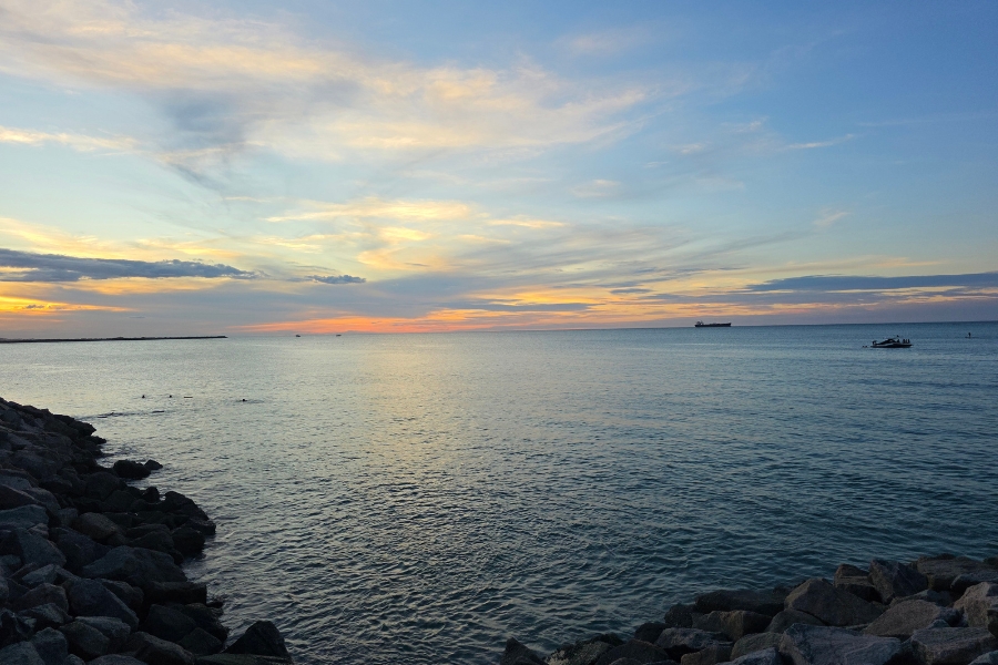 Pôr do sol dourado no mar calmo, visto de uma encosta de pedras escuras. O céu tem nuvens leves e há pequenas embarcações no horizonte.