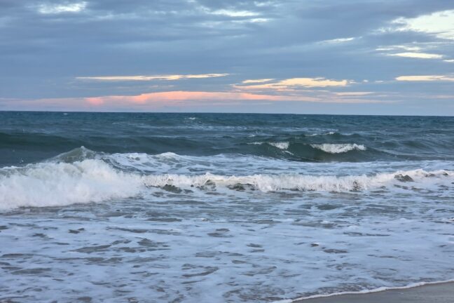 Mar com ondas quebrando em espuma branca sob um céu nublado de fim de tarde, revelando um discreto tom rosado no horizonte.