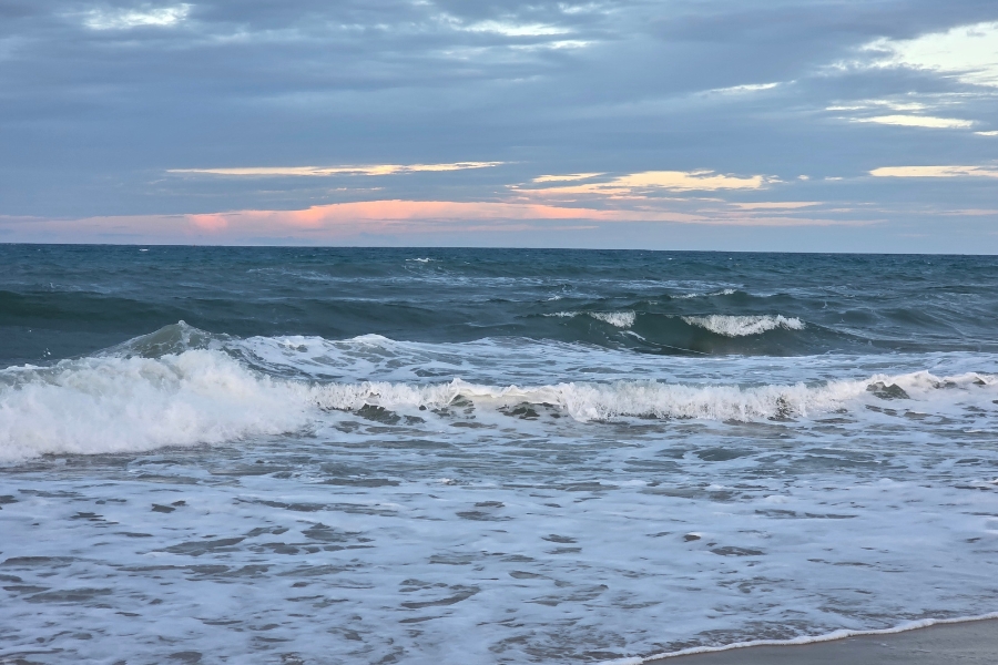 Mar com ondas quebrando em espuma branca sob um céu nublado de fim de tarde, revelando um discreto tom rosado no horizonte.