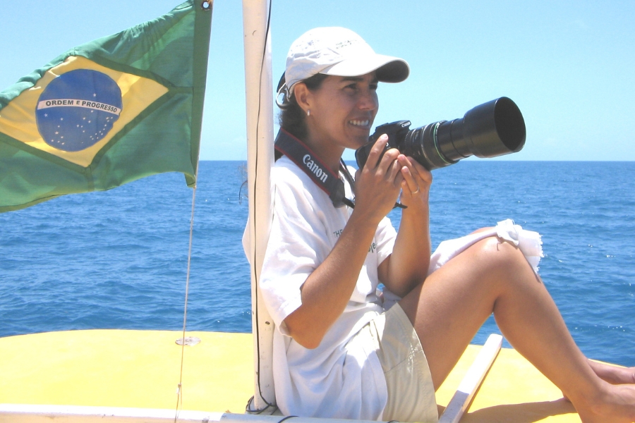 Uma mulher loira e sorridente posa de perfil em um barco no mar. Ela usa uma blusa branca e segura uma câmera fotográfica profissional com uma grande lente teleobjetiva acoplada.