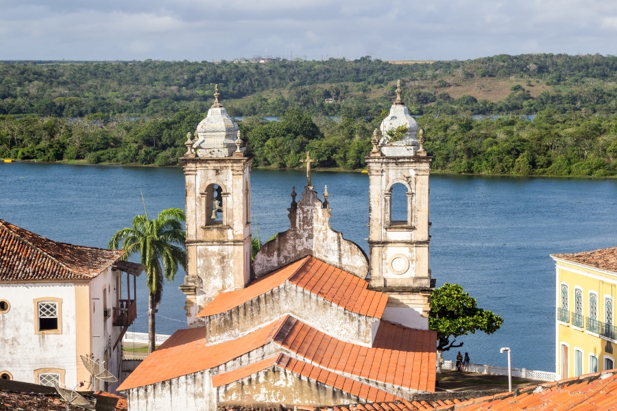 A fotografia apresenta uma vista aérea parcial de uma igreja histórica com torres brancas e telhado de barro alaranjado, situada às margens do Rio São Francisco em Alagoas. Ao fundo, as águas azuis do rio contrastam com a vegetação densa e verdejante das colinas na outra margem, sob um céu levemente nublado