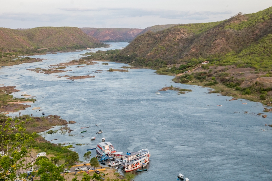 Esta vista panorâmica captura a imensidão do Rio São Francisco serpenteando entre cânions e colinas cobertas por vegetação rasteira e arbustiva. No primeiro plano, à esquerda, há um pequeno porto com embarcações de turismo atracadas, incluindo catamarãs brancos, enquanto o leito do rio revela pequenas ilhas rochosas e bancos de areia, ilustrando a geografia característica do Baixo São Francisco