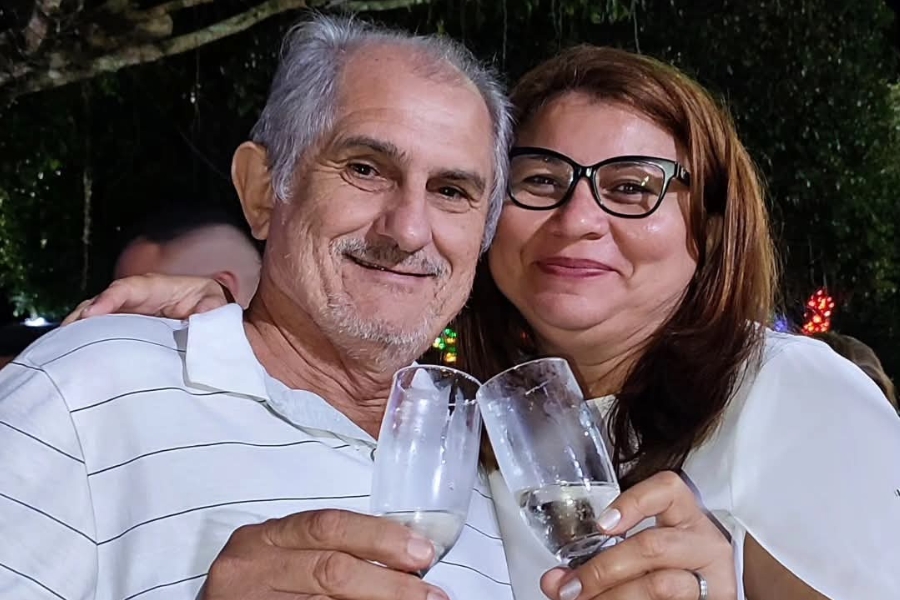 Um homem de cabelos grisalhos e camisa listrada sorri ao lado de uma mulher de óculos, enquanto ambos brindam com taças de espumante.