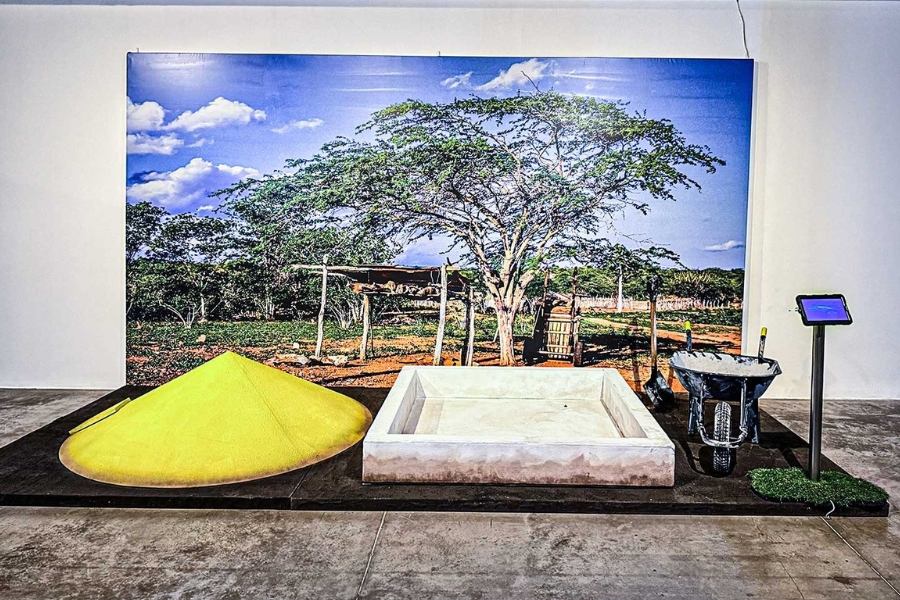 Instalação cenográfica com um monte cônico amarelo, uma estrutura quadrada de concreto, uma pá e um carrinho de mão. Ao fundo, um grande painel fotográfico retrata a vegetação do Sertão.