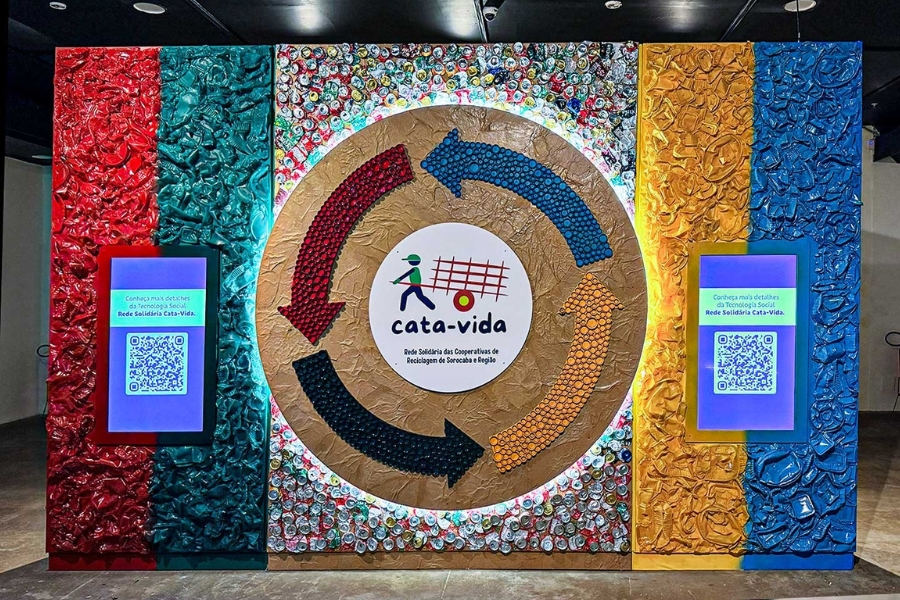Painel colorido feito com materiais reciclados. No centro, um círculo marrom com setas coloridas envolve a marca "cata-vida". Há telas digitais com QR codes nas laterais.