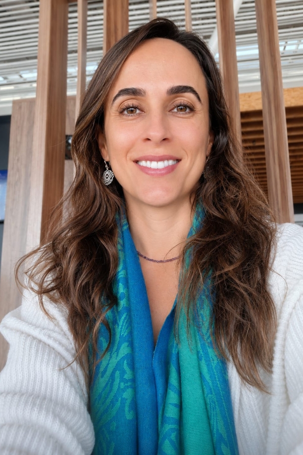 Retrato de uma mulher sorrindo, com cabelos longos ondulados, vestindo casaco branco e um lenço azul e verde estampado em volta do pescoço.