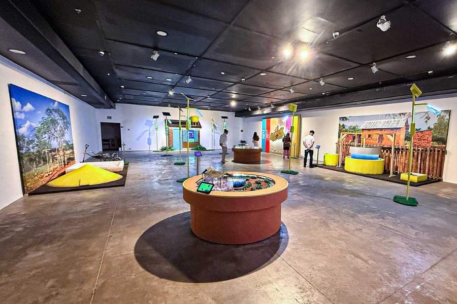 Amplo salão de exposição interna exibindo diversas maquetes e instalações interativas espalhadas pelo espaço. Algumas pessoas caminham e observam as obras sob luzes embutidas no teto.