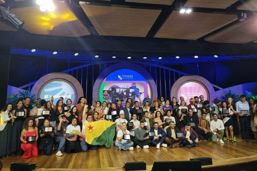 Grande grupo de pessoas reunido em um palco iluminado, muitas segurando placas de premiação. No centro, duas pessoas seguram a bandeira do estado do Acre (amarela e verde com estrela vermelha)