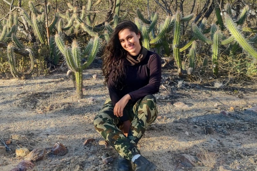 Mulher sorridente sentada no chão de terra em meio a cactos. Veste blusa preta de manga longa, calça camuflada e botas escuras.