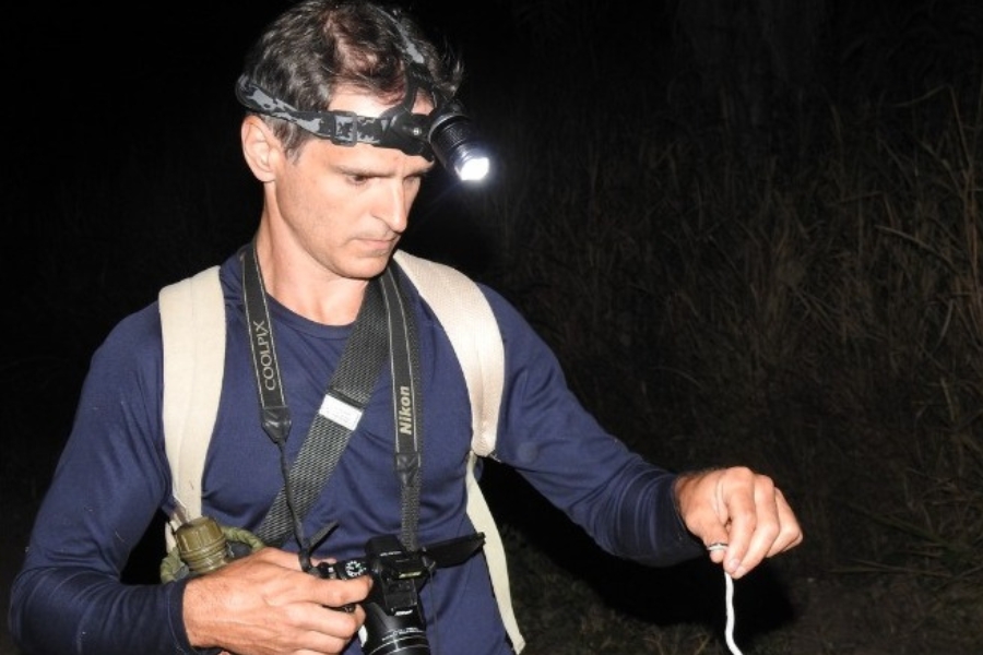 Homem à noite com lanterna de cabeça segura uma pequena cobra esbranquiçada. Ele usa mochila, camiseta azul e carrega uma câmera Nikon no pescoço.