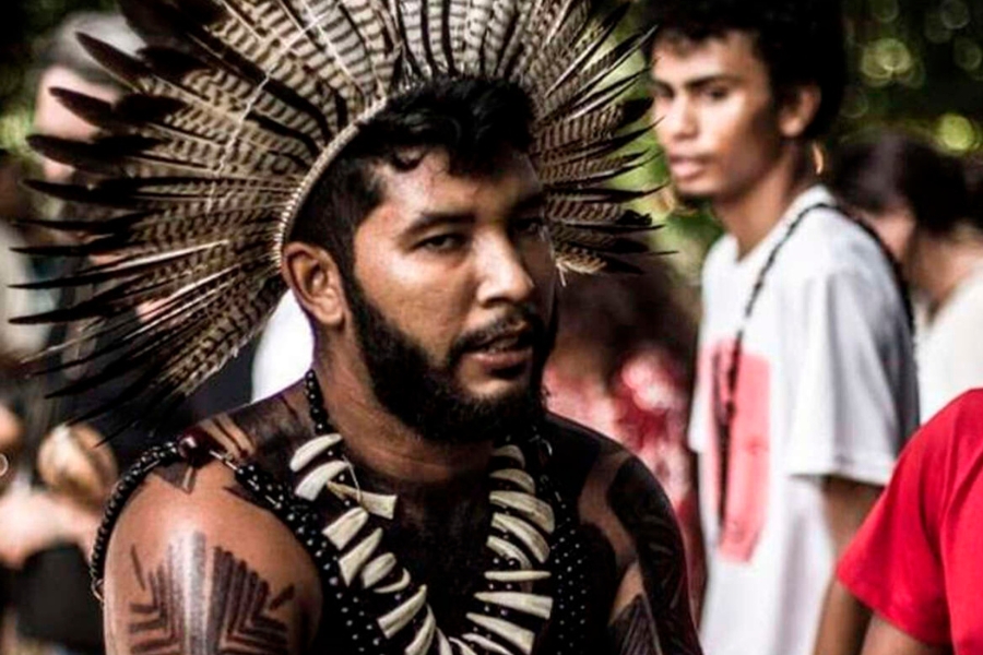 Homem indígena de barba com grande cocar de penas na cabeça e colar. Outras pessoas aparecem desfocadas ao fundo.