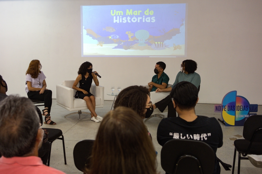 Quatro pessoas de máscara conversam em um palco durante um evento. Ao fundo, uma tela projeta a imagem "Um Mar de Histórias". Em primeiro plano, a plateia assiste sentada.