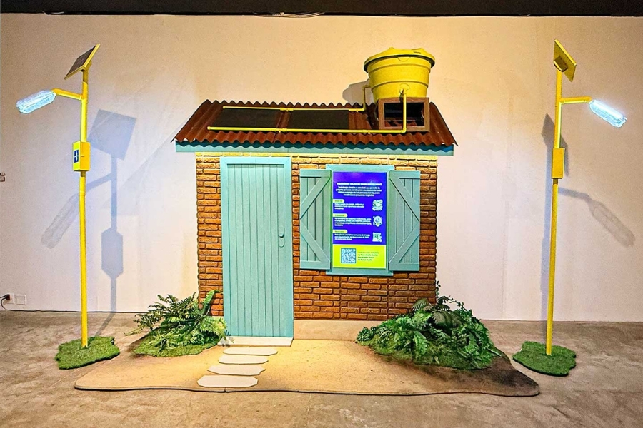 Maquete de fachada de casa de tijolos com porta e janelas azuis, painel solar e caixa-d'água amarela no telhado. Ladeada por dois postes amarelos com lâmpadas de garrafa PET.