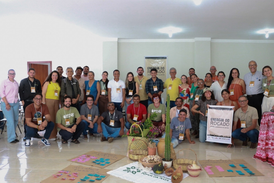 Grupo grande de pessoas posa reunido em uma sala clara, sorrindo para a foto. À frente, no chão, há cartazes com notas adesivas, cestas, cactos e elementos agrícolas.