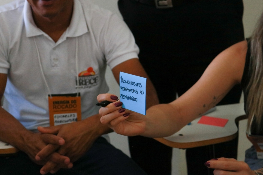 Close focado na mão de uma mulher que segura um papel adesivo azul com o texto escrito "TECNOLOGIAS ADAPTADAS AO SEMIÁRIDO". Ao fundo, aparecem outras pessoas sentadas.