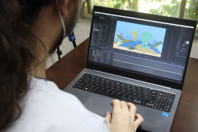 Visão por cima do ombro de uma pessoa de máscara mexendo em um notebook. Na tela do computador, há um software de edição aberto com uma animação colorida de fundo do mar.