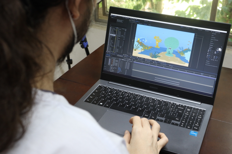 Visão por cima do ombro de uma pessoa de máscara mexendo em um notebook. Na tela do computador, há um software de edição aberto com uma animação colorida de fundo do mar.