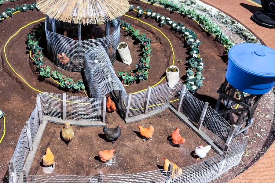 Maquete detalhada de um quintal produtivo circular com horta, galinheiro coberto de palha, cercados com galinhas em miniatura e uma caixa-d'água azul sobre suporte de madeira.
