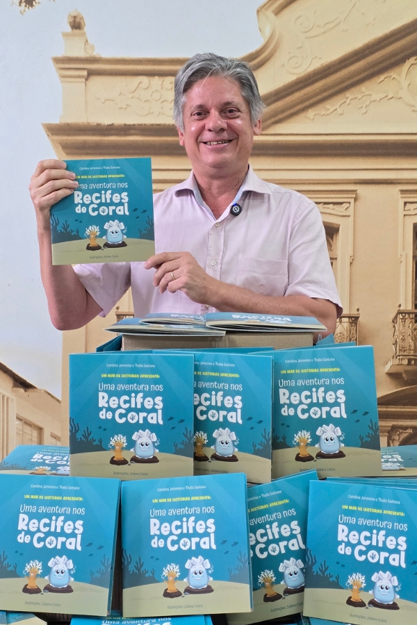 Homem sorridente de camisa clara segura um livro infantil azul intitulado "Uma aventura nos Recifes de Coral". À sua frente, há várias pilhas do mesmo livro sobre uma mesa.