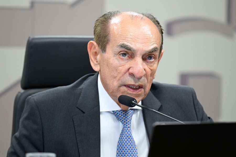 Homem de terno cinza, camisa branca e gravata azul fala ao microfone em uma mesa de reunião, posicionado em frente a uma cadeira preta de escritório.
