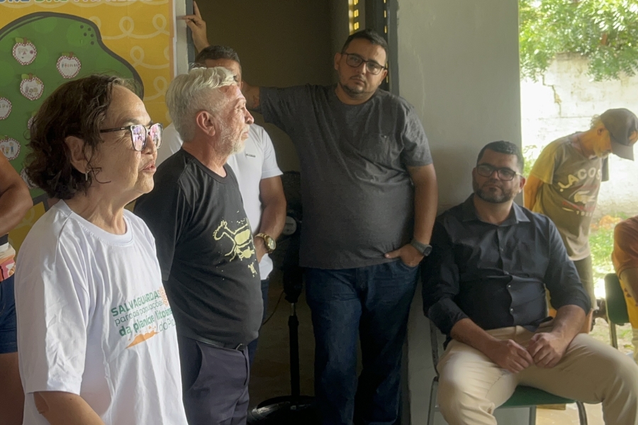 Mulher de óculos e camiseta branca fala em primeiro plano à esquerda, enquanto um grupo de cinco homens acompanha o discurso em um ambiente interno iluminado por uma porta aberta.