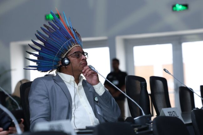 Homem de terno e cocar indígena de penas azuis e vermelhas participa de conferência internacional. Atento, ele usa fones de tradução e apoia a mão no queixo. À sua frente, há uma placa escrito "BRAZIL".