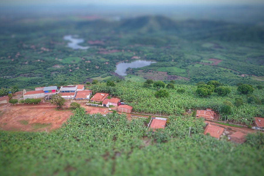 Vista aérea de um povoado cercado por plantações verdes, com casas de telhado vermelho, pequeno lago e montanhas ao fundo.