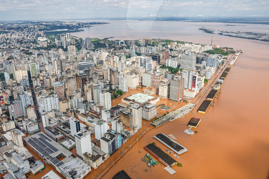 Vista aérea de uma grande cidade inundada por águas barrentas de cor marrom. A enchente encobre ruas e praças, cercando edifícios e prédios altos no centro urbano. À direita, o leito de um grande rio transbordado avança sobre as estruturas portuárias e as vias públicas da cidade.