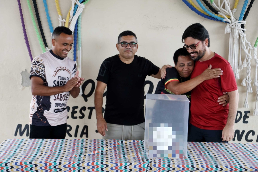 Quatro pessoas em frente a uma parede decorada: homem à esquerda aplaude e homem à direita abraça outra pessoa ao lado de caixa sobre mesa.