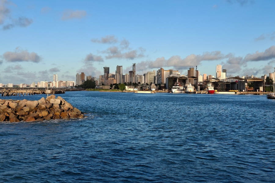 Vista panorâmica de uma orla urbana a partir do mar. Em primeiro plano, no canto esquerdo, há um espigão formado por grandes pedras sobre as águas azuis. Ao fundo, alinha-se a silhueta da cidade com diversos prédios altos, além de uma zona portuária com galpões e barcos atracados sob um céu azul com nuvens dispersas.
