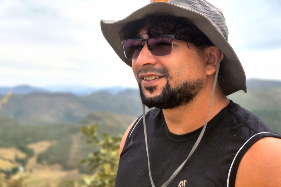 Homem sorridente com barba, óculos escuros e chapéu de canoa, usando camiseta regata preta, com paisagem de montanhas ao fundo.