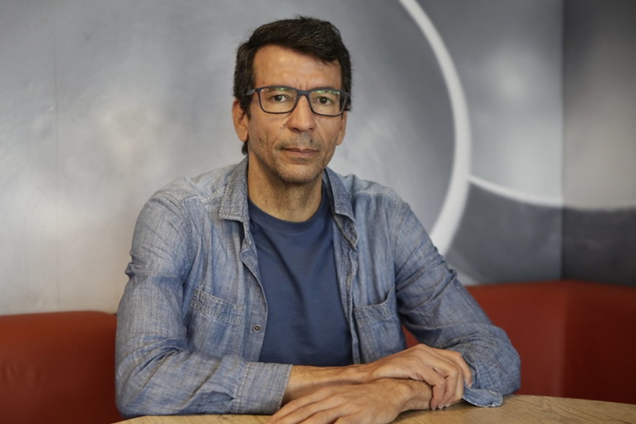 Homem de óculos e camisa jeans azul clara sobre camiseta azul-escura, sentado à mesa com as mãos cruzadas.