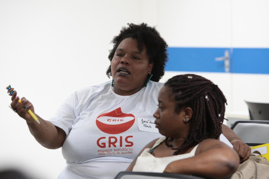 Mulher negra gesticulando ao falar durante uma reunião, vestindo camiseta branca com o logotipo "GRIS espaço solidário".