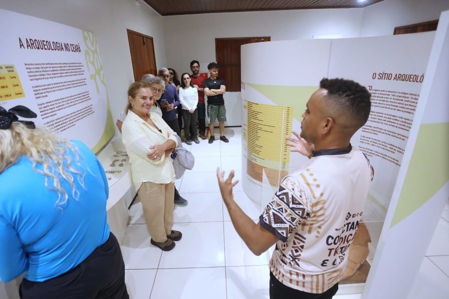 Homem de camiseta estampada explicando conteúdo de painéis explicativos sobre arqueologia para grupo de pessoas dentro de um museu.