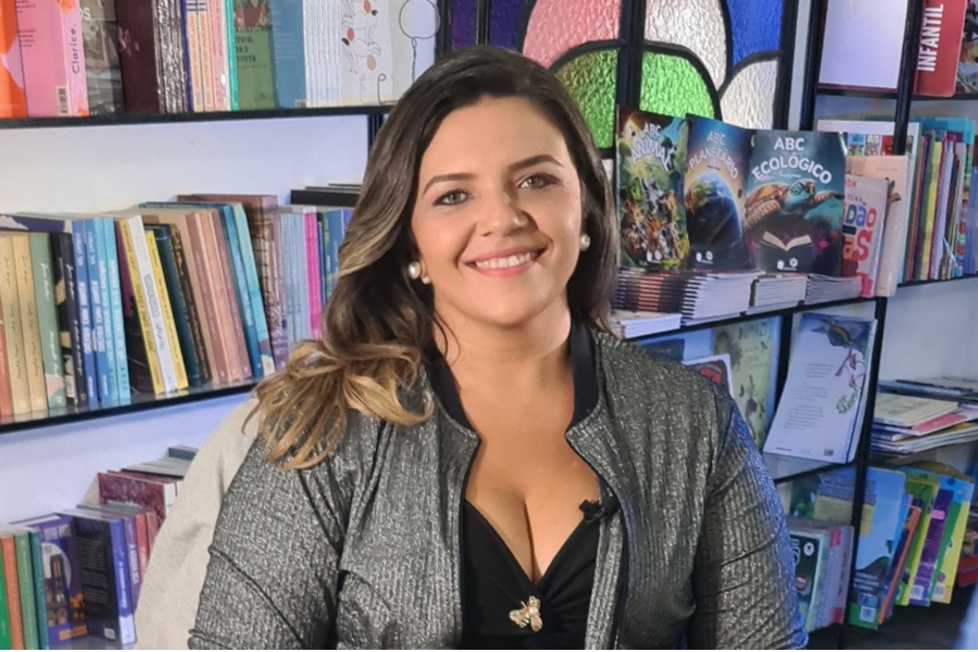 Retrato em plano médio de uma mulher sorrindo para a câmera, usando casaco cinza e blusa preta, com estantes cheias de livros ao fundo.