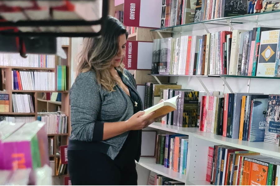 Mulher de casaco cinza em pé entre prateleiras de uma livraria, segurando um livro aberto e lendo de perfil.