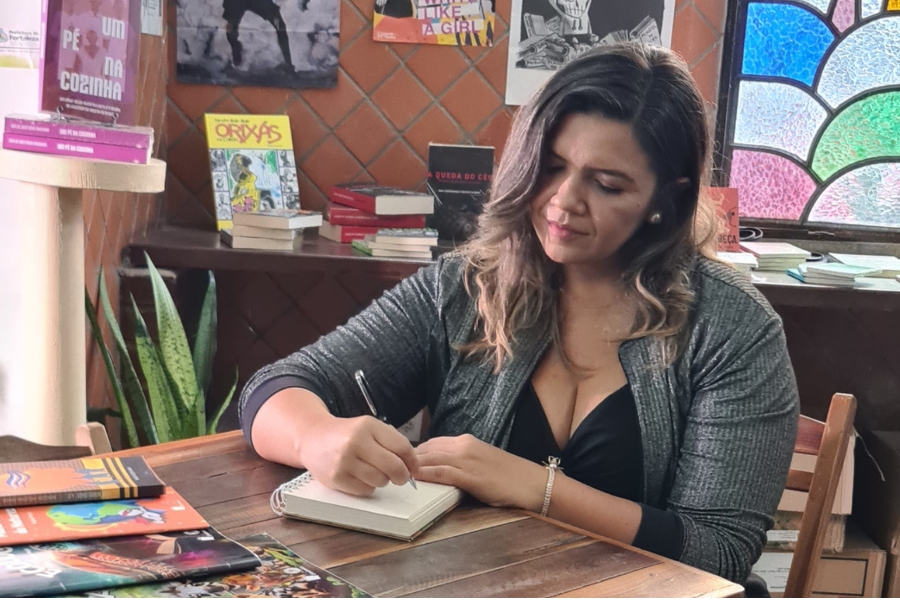 Mulher de cabelos castanhos e casaco cinza sentada à mesa de madeira, escrevendo com uma caneta em um caderno espiral. Há livros ao fundo e sobre a mesa.