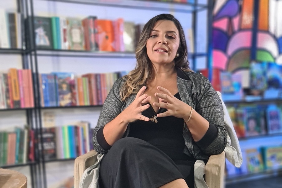 Mulher sentada em uma poltrona, falando e gesticulando com as mãos. Ela veste casaco cinza e roupa preta, diante de estantes de livros e um vitral colorido.