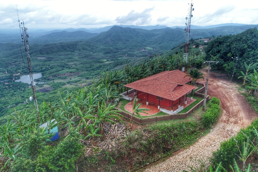 Vista aérea de casa de telhado vermelho entre bananeiras e estrada de terra, acompanhada por duas torres de comunicação e montanhas.