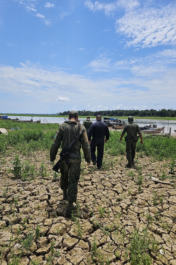 Quatro militares caminhando de costas por um terreno de solo seco e rachado em direção à margem de um rio. Os militares vestem fardas operacionais; o homem mais próximo carrega um fuzil preso ao corpo. Ao fundo, veem-se pequenas embarcações atracadas perto da margem, vegetação e um céu ensolarado com nuvens esparsas.