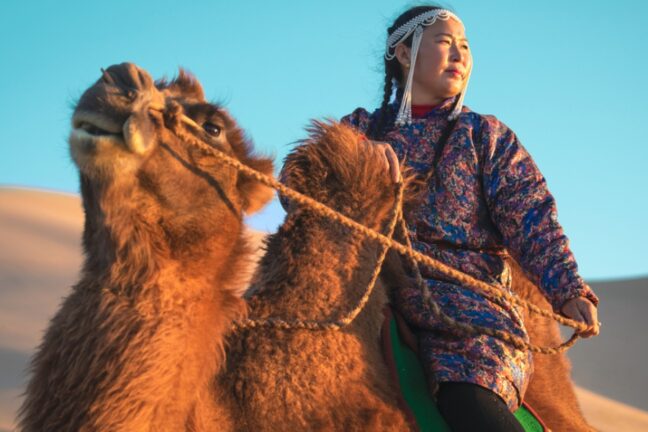 Uma mulher em trajes tradicionais mongóis e adereço na cabeça está montada em um camelo no deserto, sob a luz do sol. Ao fundo, veem-se dunas de areia e um céu azul sem nuvens.