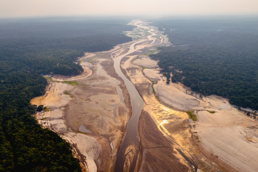 Vista aérea de um rio estreito e com grandes trechos de leito seco e exposto, cercado por uma densa floresta tropical.