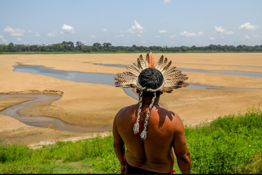 Homem indígena visto de costas usando cocar de penas, observando um leito de rio predominantemente seco com finos filetes de água.