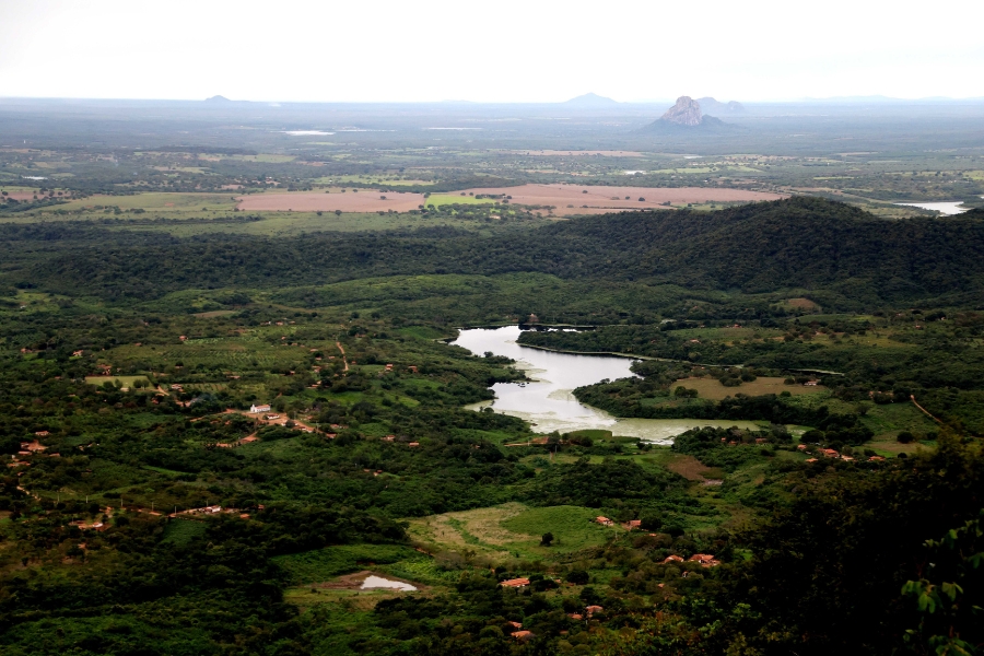 Paisagem panorâmica com extensa vegetação verde, um lago central e formações rochosas e montanhas ao fundo sob céu claro.